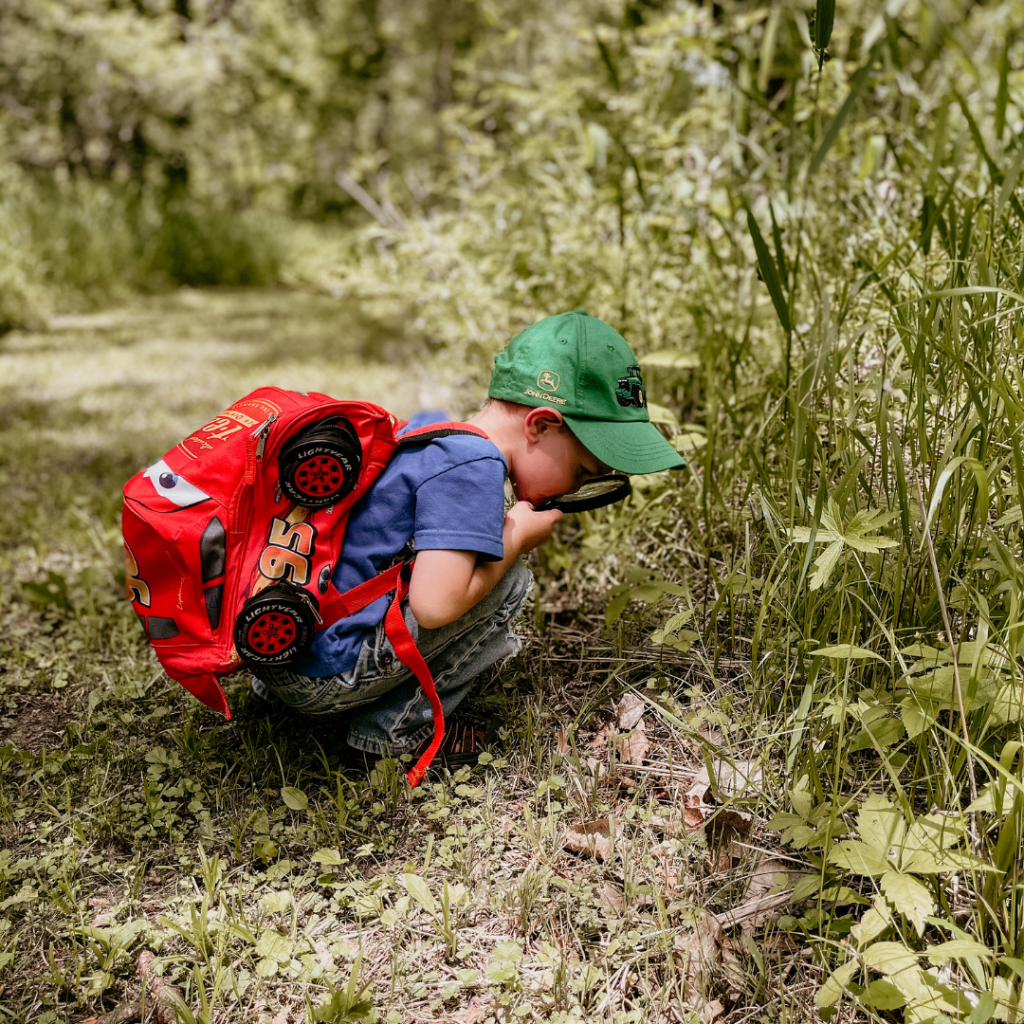 A young boy wearing a backpack looking at leaves through a magnifying glass.