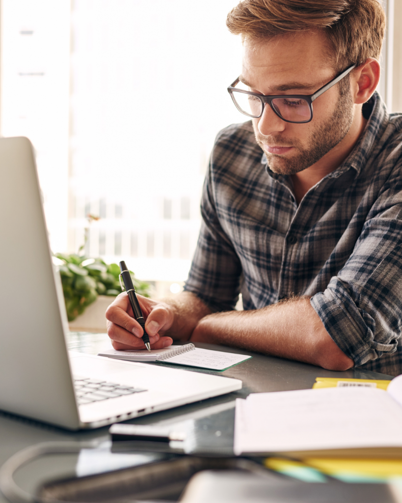 Man writing in a notebook and working a laptop in the process of working on business transition prior to full-time travel.
